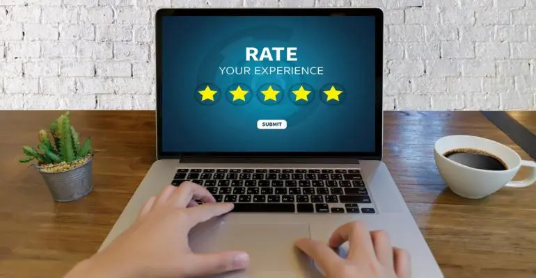 Person using a laptop displaying a "Rate Your Experience" screen with five highlighted stars, while managing storage facility software, alongside a potted cactus and a cup of coffee on a wooden table.