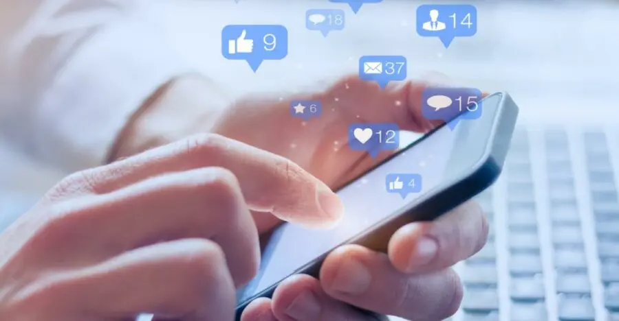 A person uses a smartphone with social media notification icons above the screen, possibly checking updates on self storage software. A laptop keyboard is visible in the background.