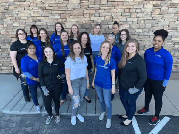A group of sixteen women stand outside on a sidewalk in front of a stone wall, most wearing blue or black company shirts, smiling at the camera—an energetic team specializing in storage facility management.