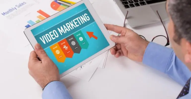 A person in business attire holds a tablet displaying a "Video Marketing" graphic with icons and an upward arrow, highlighting self storage software; sales documents are visible on the desk.