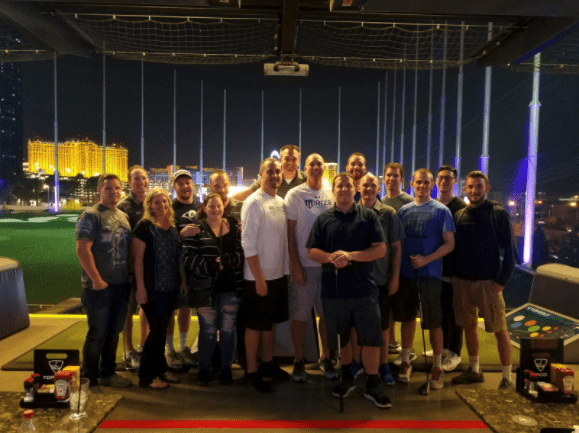 A group of people pose together at an outdoor driving range at night, with city buildings and illuminated targets visible in the background—perhaps unwinding after a long day managing a storage facility or working with self storage software.