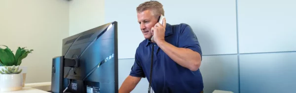 A man in a blue polo shirt stands at a desk, talking on a corded phone while looking at a computer monitor displaying self storage software.