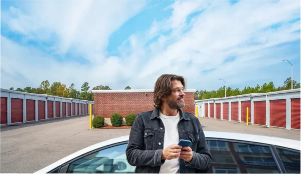 A man stands next to a white car holding a smartphone in front of a row of red storage unit doors, possibly using self storage software, under a blue sky with clouds.