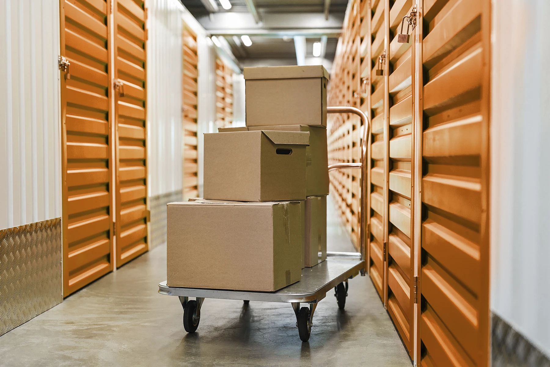 Three cardboard boxes are stacked on a flat cart in a hallway lined with orange storage unit doors, illustrating the need for efficient storage unit management.