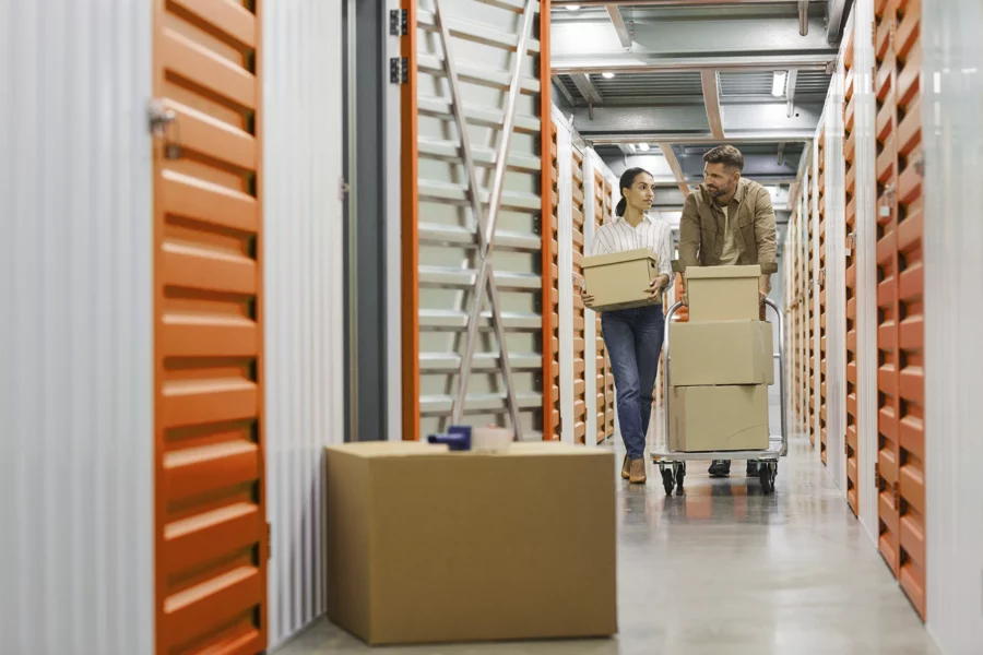 Two people move cardboard boxes through a storage facility hallway with orange and white doors, highlighting the importance of efficient storage unit management.