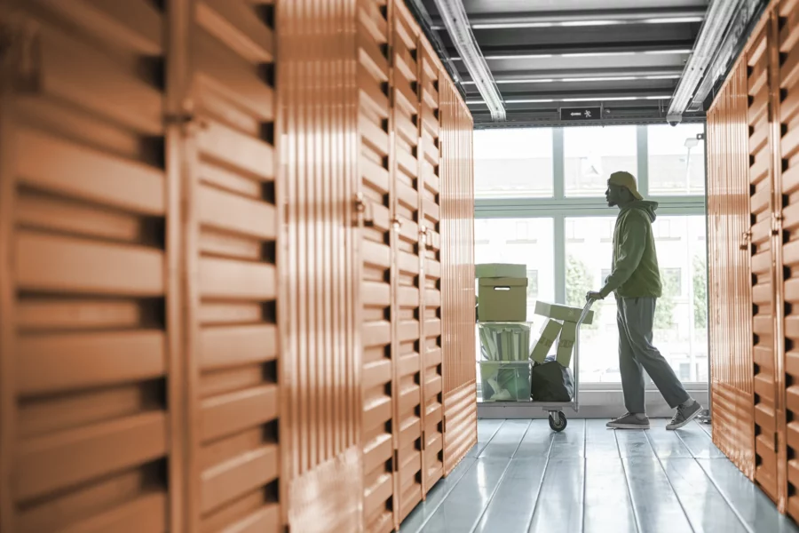 A person pushes a cart with boxes and household items down a hallway lined with orange storage unit doors, showcasing the efficiency brought by self storage software.