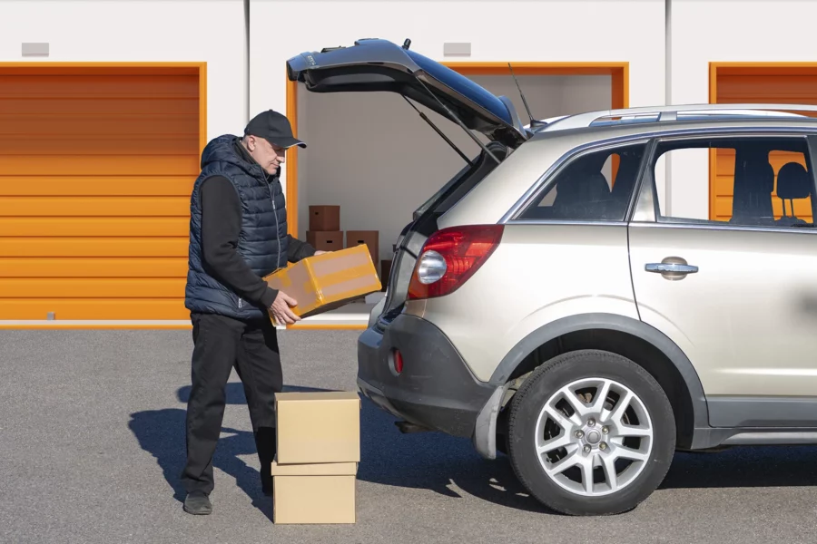 A person places a cardboard box into the trunk of an SUV parked outside orange storage unit doors on a sunny day, illustrating the convenience enabled by self storage software.