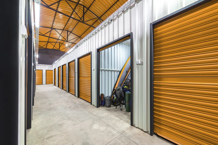 Row of indoor storage units with orange rolling doors; one unit is open, revealing assorted items such as sports equipment and boxes, ideal for those using self storage software to efficiently organize and manage their belongings.