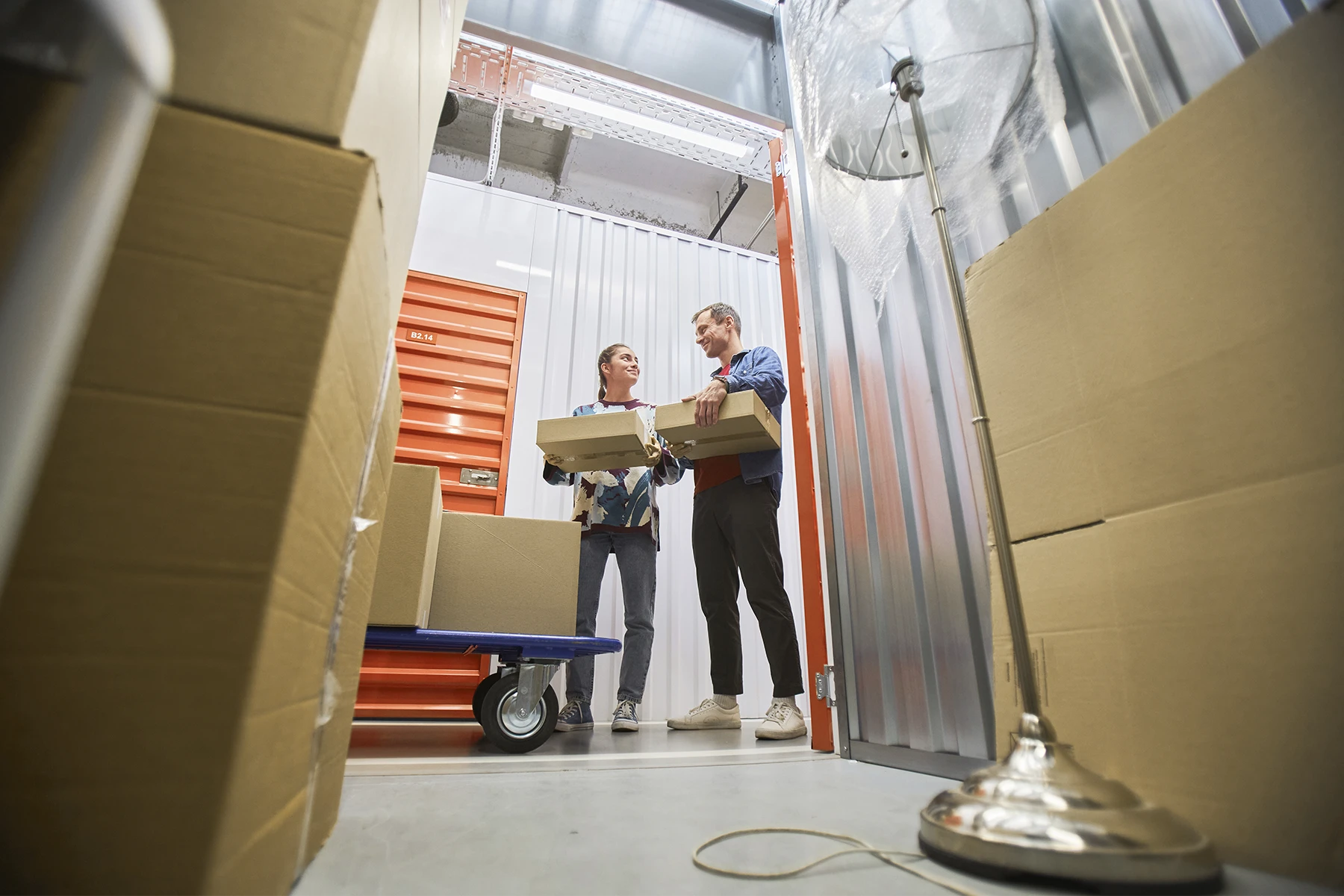 Two people stand in a storage unit holding boxes, surrounded by stacked cardboard boxes, a trolley, and a metal floor lamp—ideal for showcasing the benefits of self storage software in streamlining storage facility management.
