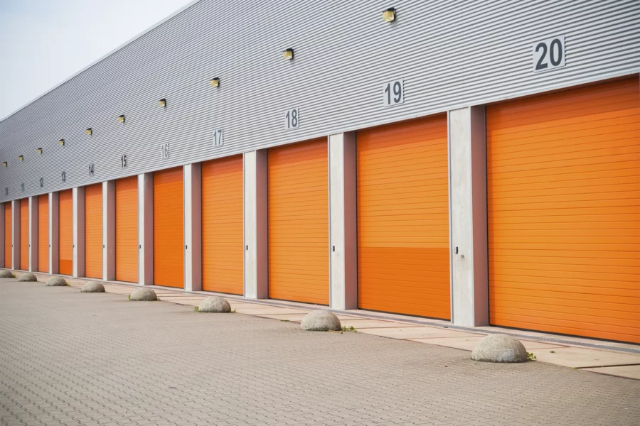 Row of orange roll-up storage unit doors on a large industrial building, numbered 16 to 20, with concrete barriers lined up in front—a practical setup for efficient storage facility management.