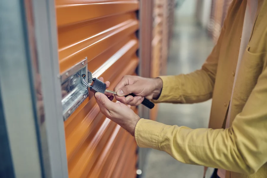 A person wearing a yellow shirt unlocks a padlock on an orange storage unit door, illustrating effective storage unit management in a hallway of storage lockers.