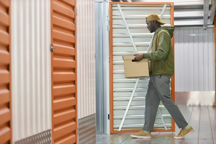 A person in casual clothing carries a cardboard box into an open storage unit with orange and white doors in a brightly lit facility, showcasing efficient storage unit management.