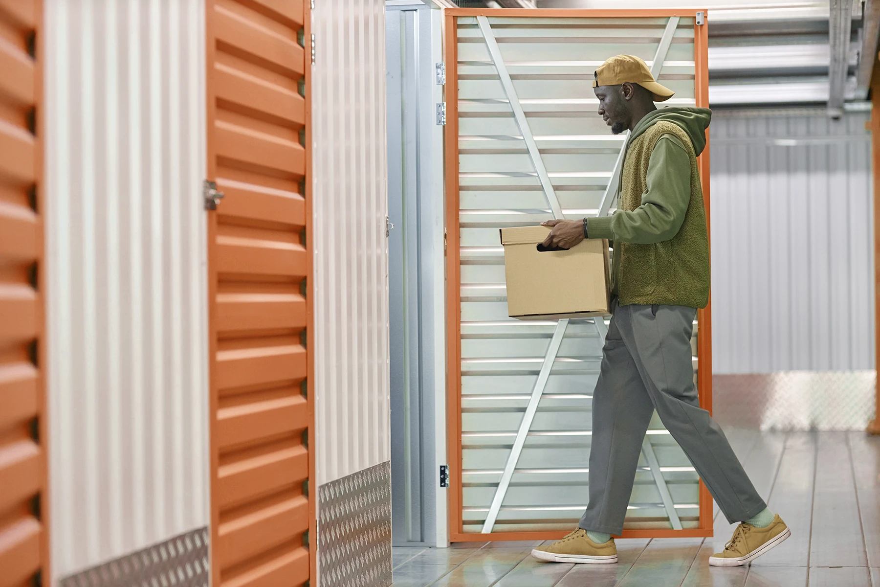 A person in casual clothing carries a cardboard box into an open storage unit with orange and white doors in a brightly lit facility, showcasing efficient storage unit management.
