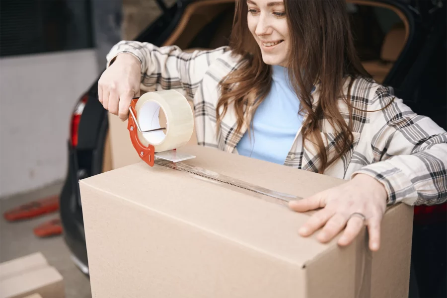 A woman seals a cardboard box with packing tape near the open trunk of a car, streamlining her move with the help of self storage software.